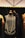 A vintage-style black dress displayed on a mannequin with a long pearl necklace draped around it. The dress features intricate beading and fringe details. It is surrounded by other mannequins wearing elegant garments, and the background consists of a dark curtain and a geometric ceiling design.