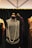 A vintage-style black dress displayed on a mannequin with a long pearl necklace draped around it. The dress features intricate beading and fringe details. It is surrounded by other mannequins wearing elegant garments, and the background consists of a dark curtain and a geometric ceiling design.
