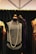 A vintage-style black dress displayed on a mannequin with a long pearl necklace draped around it. The dress features intricate beading and fringe details. It is surrounded by other mannequins wearing elegant garments, and the background consists of a dark curtain and a geometric ceiling design.