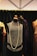 A vintage-style black dress displayed on a mannequin with a long pearl necklace draped around it. The dress features intricate beading and fringe details. It is surrounded by other mannequins wearing elegant garments, and the background consists of a dark curtain and a geometric ceiling design.