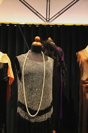 A vintage-style black dress displayed on a mannequin with a long pearl necklace draped around it. The dress features intricate beading and fringe details. It is surrounded by other mannequins wearing elegant garments, and the background consists of a dark curtain and a geometric ceiling design.