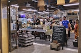 A bustling Italian market with customers browsing and purchasing items. The interior is decorated with hanging wicker baskets and shelves filled with various products, including tins and jars. There is a chalkboard sign advertising special offers, and the atmosphere is lively and vibrant.