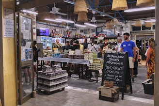 A bustling Italian market with customers browsing and purchasing items. The interior is decorated with hanging wicker baskets and shelves filled with various products, including tins and jars. There is a chalkboard sign advertising special offers, and the atmosphere is lively and vibrant.