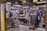A bustling Italian market with customers browsing and purchasing items. The interior is decorated with hanging wicker baskets and shelves filled with various products, including tins and jars. There is a chalkboard sign advertising special offers, and the atmosphere is lively and vibrant.