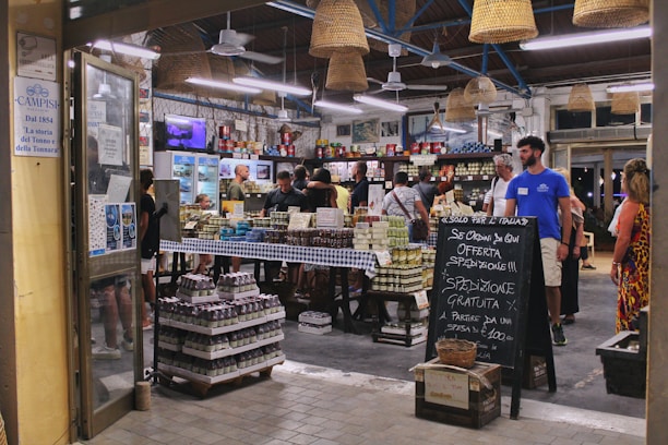 A bustling Italian market with customers browsing and purchasing items. The interior is decorated with hanging wicker baskets and shelves filled with various products, including tins and jars. There is a chalkboard sign advertising special offers, and the atmosphere is lively and vibrant.
