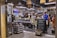 A bustling Italian market with customers browsing and purchasing items. The interior is decorated with hanging wicker baskets and shelves filled with various products, including tins and jars. There is a chalkboard sign advertising special offers, and the atmosphere is lively and vibrant.