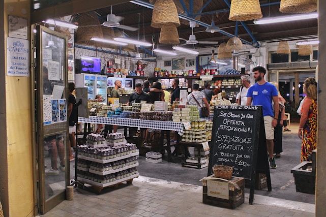 A bustling Italian market with customers browsing and purchasing items. The interior is decorated with hanging wicker baskets and shelves filled with various products, including tins and jars. There is a chalkboard sign advertising special offers, and the atmosphere is lively and vibrant.