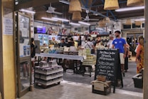 A bustling Italian market with customers browsing and purchasing items. The interior is decorated with hanging wicker baskets and shelves filled with various products, including tins and jars. There is a chalkboard sign advertising special offers, and the atmosphere is lively and vibrant.