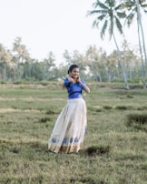 A woman stands in a grassy field with palm trees in the background. She is wearing a traditional outfit consisting of a blue blouse and a long beige skirt adorned with blue motifs. Her hair is styled back, and she is smiling while adjusting her earrings.