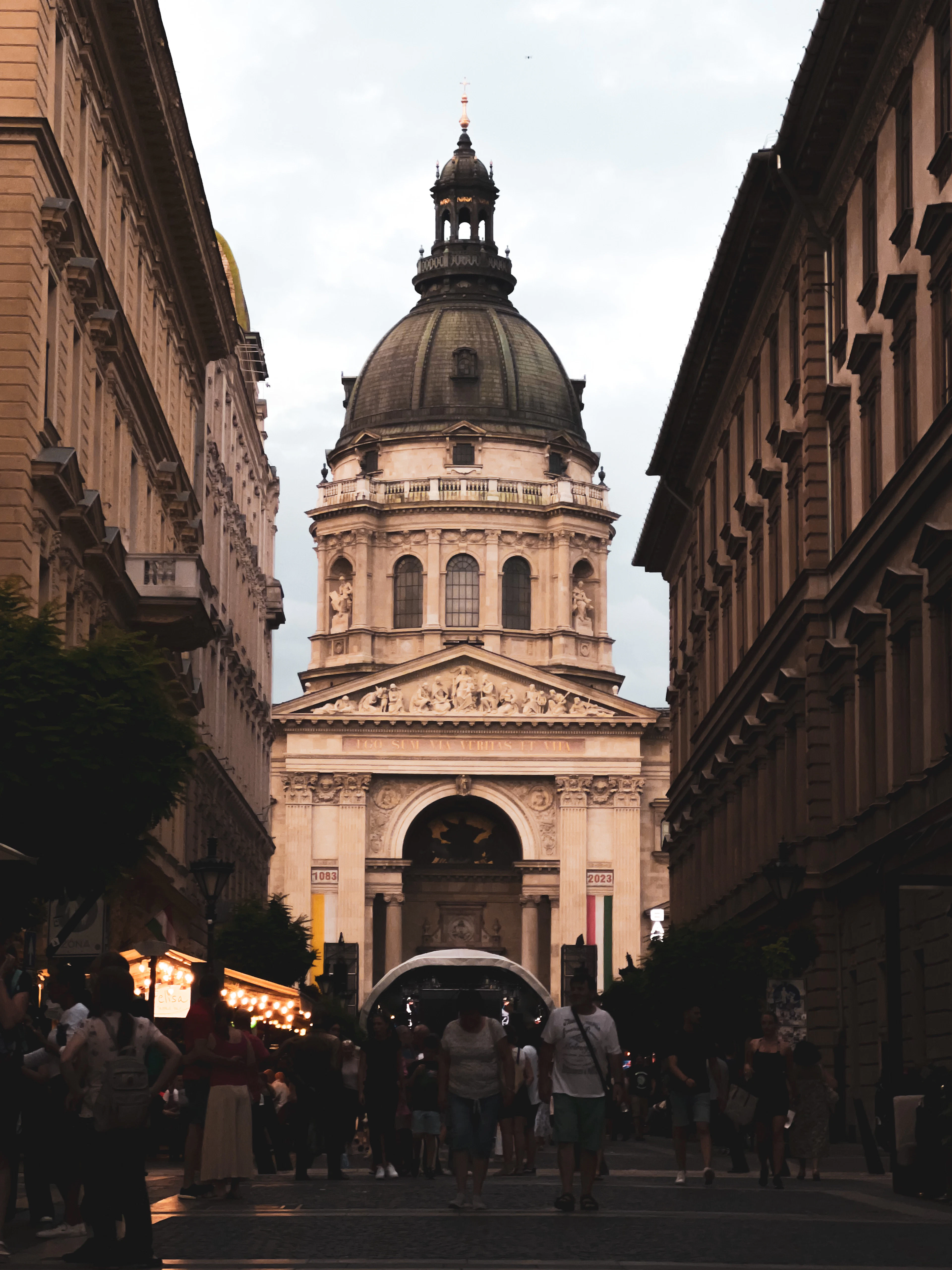 The green dome and columned portico of St Stephen's Basilica in Budapest closing the view down a pedestrian street of ochre apartment blocks, with people walking towards it in soft evening light