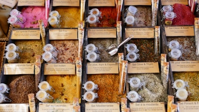 An array of colorful and diverse spices in individual compartments, each labeled with descriptions in French. The vibrant spices range from pink, red, and orange to yellow and brown, each accompanied by small glass jars. A metal scoop is placed among the spices.