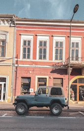 A vintage off-road vehicle is parked in front of a historic European building with red and beige walls. The building features large windows with white trim, and there is a streetlamp on the right. The car has a blue body with a black roof and is parked on a cobblestone pavement.