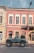A vintage off-road vehicle is parked in front of a historic European building with red and beige walls. The building features large windows with white trim, and there is a streetlamp on the right. The car has a blue body with a black roof and is parked on a cobblestone pavement.