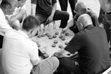 A group of men are gathered around a small square table, deeply engaged in a game. The game involves circular pieces with characters, arranged on a grid. The setting suggests they are playing a traditional board game, with intense focus and strategic thinking apparent from their expressions and body language. The image is in black and white, capturing a timeless, classic feel.