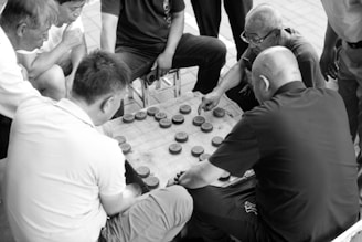 A group of men are gathered around a small square table, deeply engaged in a game. The game involves circular pieces with characters, arranged on a grid. The setting suggests they are playing a traditional board game, with intense focus and strategic thinking apparent from their expressions and body language. The image is in black and white, capturing a timeless, classic feel.