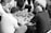 A group of men are gathered around a small square table, deeply engaged in a game. The game involves circular pieces with characters, arranged on a grid. The setting suggests they are playing a traditional board game, with intense focus and strategic thinking apparent from their expressions and body language. The image is in black and white, capturing a timeless, classic feel.