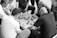 A group of men are gathered around a small square table, deeply engaged in a game. The game involves circular pieces with characters, arranged on a grid. The setting suggests they are playing a traditional board game, with intense focus and strategic thinking apparent from their expressions and body language. The image is in black and white, capturing a timeless, classic feel.