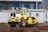 A yellow steamroller is parked on a construction site, surrounded by dirt and earth. In the background, there are portable white containers and storage buildings with blue doors.