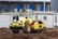 A yellow steamroller is parked on a construction site, surrounded by dirt and earth. In the background, there are portable white containers and storage buildings with blue doors.