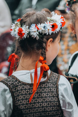 A young girl with braided hair adorned with a vibrant floral crown, featuring red and white flowers. She wears a patterned vest over a white shirt, tied at the back with orange ribbons. The focus is on the back of her head, emphasizing the intricate details of her attire and hairstyle.
