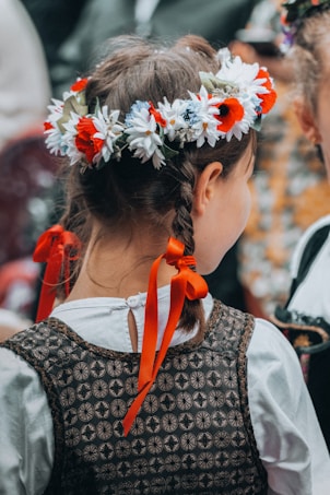 A young girl with braided hair adorned with a vibrant floral crown, featuring red and white flowers. She wears a patterned vest over a white shirt, tied at the back with orange ribbons. The focus is on the back of her head, emphasizing the intricate details of her attire and hairstyle.