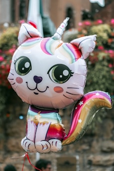 A colorful balloon shaped like a cute cat-unicorn hybrid, featuring large expressive eyes and a rainbow-colored mane and tail. The balloon is set against an outdoor background with blurred, natural elements.