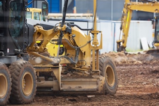 Several pieces of construction machinery are present on a dirt-filled construction site. The focus is on a large, yellow bulldozer with visible tracks and hydraulic components. In the background, other construction equipment, such as an excavator, can be seen, along with a blue building and temporary fencing.