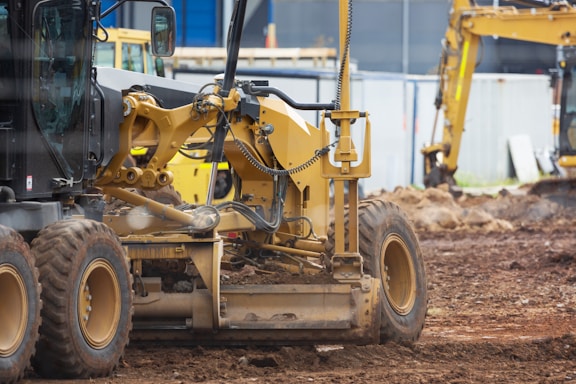 Several pieces of construction machinery are present on a dirt-filled construction site. The focus is on a large, yellow bulldozer with visible tracks and hydraulic components. In the background, other construction equipment, such as an excavator, can be seen, along with a blue building and temporary fencing.