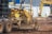 Several pieces of construction machinery are present on a dirt-filled construction site. The focus is on a large, yellow bulldozer with visible tracks and hydraulic components. In the background, other construction equipment, such as an excavator, can be seen, along with a blue building and temporary fencing.