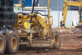 Several pieces of construction machinery are present on a dirt-filled construction site. The focus is on a large, yellow bulldozer with visible tracks and hydraulic components. In the background, other construction equipment, such as an excavator, can be seen, along with a blue building and temporary fencing.