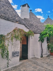 A traditional stone building with conical roofs, characteristic of Trullo architecture, featuring lush greenery climbing the walls and a sign next to a dark wooden door. The sky is bright blue with scattered clouds.