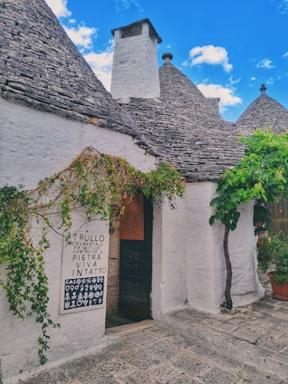 A traditional stone building with conical roofs, characteristic of Trullo architecture, featuring lush greenery climbing the walls and a sign next to a dark wooden door. The sky is bright blue with scattered clouds.