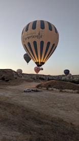 A serene landscape featuring several hot air balloons in the sky over a rocky terrain. The largest balloon in the foreground has the text 'Connecting Cultures Euroriente Travel,' and smaller balloons are visible in the background. The scene appears to be taken during early morning or late afternoon, as the lighting suggests a golden hour ambiance. Below, a vehicle and a small group of people can be seen on the ground.