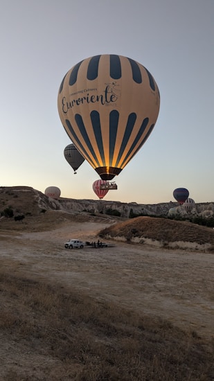 A serene landscape featuring several hot air balloons in the sky over a rocky terrain. The largest balloon in the foreground has the text 'Connecting Cultures Euroriente Travel,' and smaller balloons are visible in the background. The scene appears to be taken during early morning or late afternoon, as the lighting suggests a golden hour ambiance. Below, a vehicle and a small group of people can be seen on the ground.