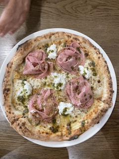 A wood-fired pizza topped with slices of pink meat, possibly ham or prosciutto, dollops of white cheese, and green herb dressing. The crust is golden brown with slightly charred edges. A hand is seen reaching towards the top of the pizza.