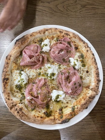 A wood-fired pizza topped with slices of pink meat, possibly ham or prosciutto, dollops of white cheese, and green herb dressing. The crust is golden brown with slightly charred edges. A hand is seen reaching towards the top of the pizza.