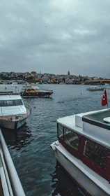 Boats are docked along a waterfront with a view of a city skyline in the background. The sky is overcast, and the water appears calm. A prominent tower is visible among the city buildings. A Turkish flag is seen on one of the boats.