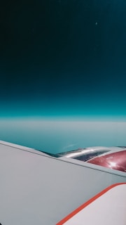 An aerial view showing an airplane wing and engine against a backdrop of the sky. The horizon is visible in the distance, with a gradient from deep blue at the top to lighter shades near the horizon.