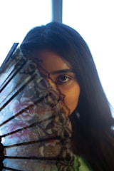 A person with long dark hair is partially concealed behind a decorative, floral-patterned hand fan. The focus is on one eye, creating a mysterious and intimate mood. The background is blurred, emphasizing the subject.