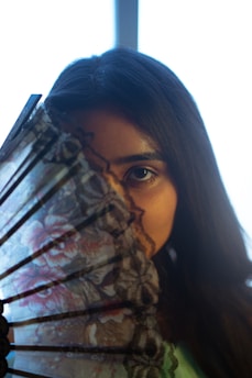 A person with long dark hair is partially concealed behind a decorative, floral-patterned hand fan. The focus is on one eye, creating a mysterious and intimate mood. The background is blurred, emphasizing the subject.