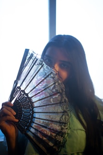A person with long dark hair partially hides their face behind an intricate lace fan. Soft lighting from a window creates a silhouette effect, highlighting the delicate patterns of the fan and casting gentle shadows.