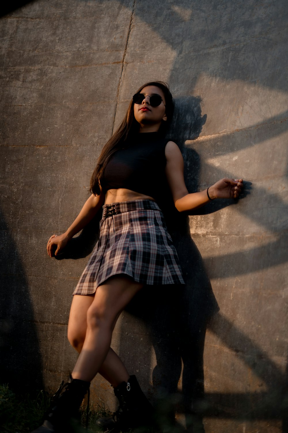 a woman wearing a skirt and sunglasses leaning against a wall