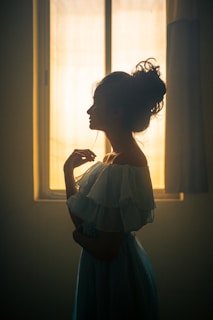 An elegant woman in a minimalist yet romantic dress standing by a window with soft natural light highlighting her silhouette.