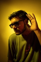 A young man is wearing dark sunglasses and a t-shirt with a graphic design. He holds his right hand up to his ear, as if trying to listen intently. The lighting casts a yellowish tint over the entire image, creating a dramatic effect.