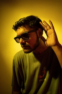 A young man is wearing dark sunglasses and a t-shirt with a graphic design. He holds his right hand up to his ear, as if trying to listen intently. The lighting casts a yellowish tint over the entire image, creating a dramatic effect.