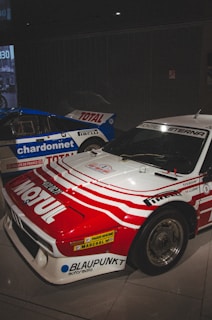 Two race cars are positioned next to each other indoors. The car in the foreground has a red and white color scheme with various sponsor logos, including Motul and Blaupunkt, prominently displayed. The car in the background is blue and white, with large logos of Total and Chardonnet. Both cars have sleek, aerodynamic designs typical of rally or racing vehicles, and the setting appears to be a showroom or museum with dim lighting.