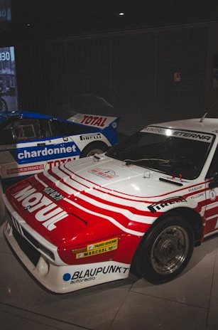 Two race cars are positioned next to each other indoors. The car in the foreground has a red and white color scheme with various sponsor logos, including Motul and Blaupunkt, prominently displayed. The car in the background is blue and white, with large logos of Total and Chardonnet. Both cars have sleek, aerodynamic designs typical of rally or racing vehicles, and the setting appears to be a showroom or museum with dim lighting.