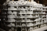 A highly detailed architectural model of an intricate building featuring curved lines and organic shapes. The facade includes multiple floors with ornate balcony railings that display complex, artistic designs. The overall texture is rough with a monochromatic color scheme, emphasizing the building's unique and fluid structure.
