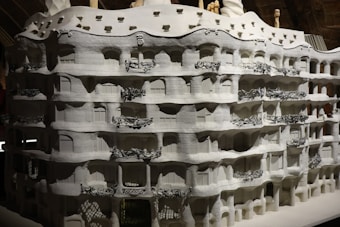 A highly detailed architectural model of an intricate building featuring curved lines and organic shapes. The facade includes multiple floors with ornate balcony railings that display complex, artistic designs. The overall texture is rough with a monochromatic color scheme, emphasizing the building's unique and fluid structure.