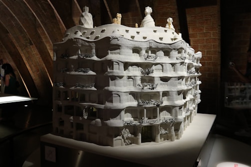 A detailed architectural model of a building with a wavy facade, multiple balconies, and elaborate decorative elements sits under a softly lit environment. The model is monochromatic, primarily white, and features intricate designs that highlight its artistic and architectural features. The background consists of curved, exposed brickwork adding texture and depth.